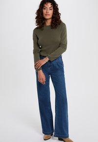 Olijfgroene lange mouw top met gegolfde halslijn, gecombineerd met hoog-getailleerde spijkerbroek met wijde pijpen in blauw. Bruine schoenen maken de outfit af.