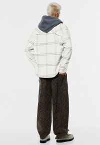 Nadměrně bílá kostkovaná bunda se šedou kapucí, hnědé vzorované kalhoty se širokými nohavicemi a béžová texturovaná obuv; viděno ze zad.