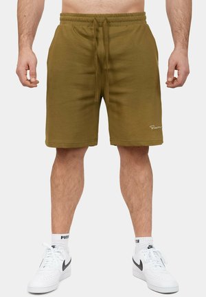 Mann trägt olivgrüne Shorts mit Kordelzug und weiße Nike-Sneaker mit Puma-Socken, steht vor einem einfarbigen hellen Hintergrund.