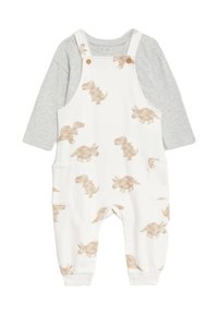 Weiße Latzhose mit beigen Dinosaurier-Prints, darunter ein graues Langarmshirt. Hergestellt aus weicher Baumwolle, mit seitlichen Taschen und Knöpfen.