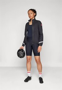 Moteris juodais dviračių drabužiais ir striuke laiko juodą šalmą, vilkinti baltas kojines su raudonomis juostelėmis ir juodus dviračių batus.