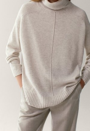 Femme portant un pull col roulé beige clair avec une couture centrale et un pantalon ample assorti, les mains dans les poches, debout devant un fond neutre.