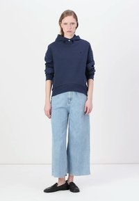 Felpa con cappuccio blu navy e colletto con bottoni, abbinata a jeans azzurri a gamba larga e scarpe nere slip-on, in piedi su uno sfondo bianco.