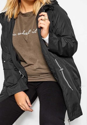 Kvinde iført sort jakke over brun skjorte med hvid tekst, sorte bukser, og holder jakke kraven med den ene hånd.