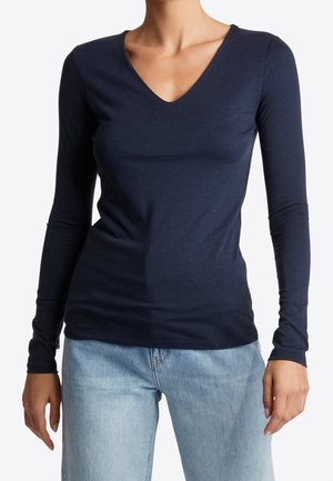 Frau trägt ein eng anliegendes, marineblaues Langarmshirt mit V-Ausschnitt und hellblaue Jeans, steht vor einem schlichten weißen Hintergrund.