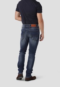 Jeans de mezclilla azul oscuro con corte ajustado, con un parche de cuero marrón en la cintura y un leve desgastado en los muslos. Zapatos visibles en la parte inferior.