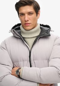 Chaqueta acolchada en gris claro con forro y capucha negra, combinada con un suéter de cuello alto grueso en beige. Reloj con correa negra visible.