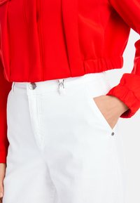 Red cropped jacket with gathered hem, paired with white pants featuring a button and loop closure, side pockets, and a smooth texture.