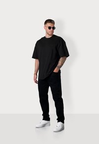 Black oversized cotton t-shirt, black pants, white sneakers; model with tattoos wearing dark sunglasses stands against a plain background.