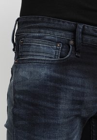 Mörkblå denimjeans med slim fit, med flera fickor, kopparnitar och en subtil blekt textur på tyget.