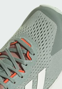 Scarpa da ginnastica grigia chiara con tomaia in mesh testurizzato, dettagli arancioni e lacci grigi. Presenta tre strisce bianche e una punta rinforzata.