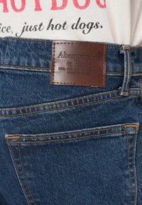 Κοντινό πλάνο μπλε τζιν με καφέ δερμάτινο ταμπελάκι Abercrombie & Fitch πάνω από την πίσω τσέπη, φορεμένα με λευκό πουκάμισο εκτυπωμένο με τη φράση «just hot dogs».