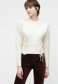 Uma mulher a usar um suéter creme com detalhes lateral em atacadores e calças castanhas escuras de cintura alta, de pé contra um fundo branco plain.