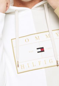 Mani che sistemano il cappuccio su una felpa bianca con logo rettangolare bordato d'oro che mostra "Tommy Hilfiger" e una piccola bandiera rossa, bianca e blu.