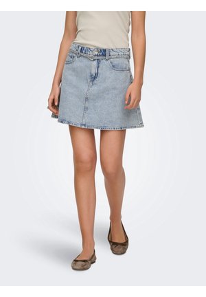 Femme portant une jupe en jean bleu clair avec une taille ceinturée, un haut beige et des ballerines marron, debout sur un fond blanc.