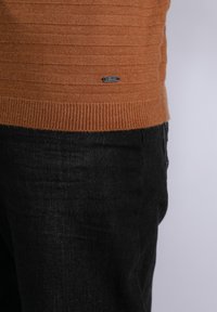 Maglione arancione lavorato a maglia con texture a coste orizzontali e un'etichetta metallica nera sul lato inferiore, abbinato a pantaloni in denim neri.