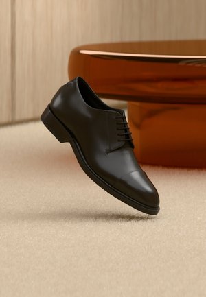 Scarpa elegante da uomo in pelle nera con lacci che sembra fluttuare sopra un tappeto beige, con un tavolo rotondo arancione sullo sfondo.