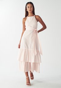 Vestido midi a rayas en rosa claro y blanco, con cuello halter, cinturilla y dobladillo de volantes en capas. Usado con sandalias blancas de tiras en el tobillo.