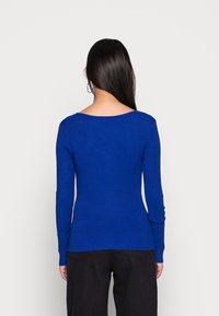 Frau mit langen dunklen Haaren, die einen figurbetonten, langärmeligen, königsblauen Rippensweater und weite schwarze Hosen trägt, steht mit dem Rücken zur Kamera.