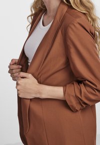 Un blazer marron à coupe décontractée, avec des manches longues et une texture lisse, partiellement ouvert pour révéler un haut de couleur claire en dessous.