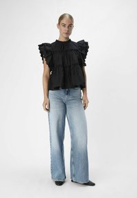 Schwarze rüschierte Bluse mit gestuftem Design, hohem Kragen und kurzen Puffärmeln, kombiniert mit hellblauen, weit geschnittenen Jeans und schwarzem Schuhwerk.