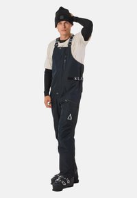 Marineblauwe overalls van duurzame stof, voorzien van ritsen, verstelbare banden en logo accenten. Gecombineerd met een zwarte beanie en zwarte laarzen.