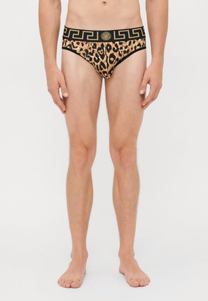 Mann, der barfuß steht und Leopardenmuster-Unterhosen mit einem schwarz-goldenen gemusterten Bund trägt, vor einem weißen Hintergrund.