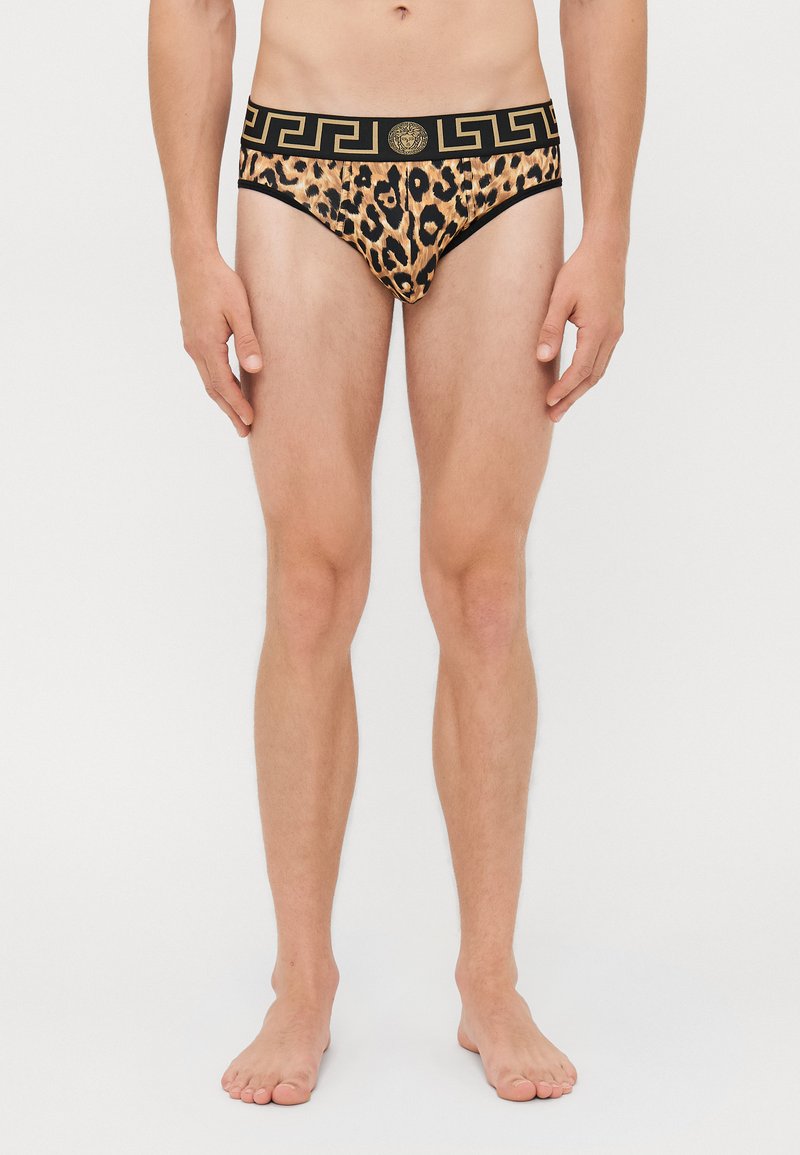 Mann, der barfuß steht und Leopardenmuster-Unterhosen mit einem schwarz-goldenen gemusterten Bund trägt, vor einem weißen Hintergrund.