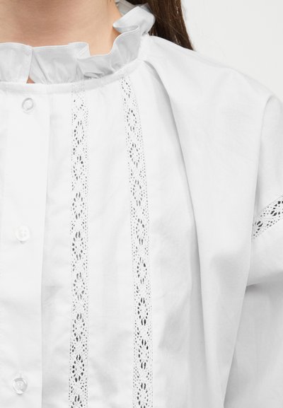 Λευκό πουκάμισο με γιακά με βολάν και κάθετες λεπτομέρειες από δαντέλα, μπροστινό κούμπωμα με κουμπιά, φορεμένο από άτομο με μακριά σκούρα μαλλιά.