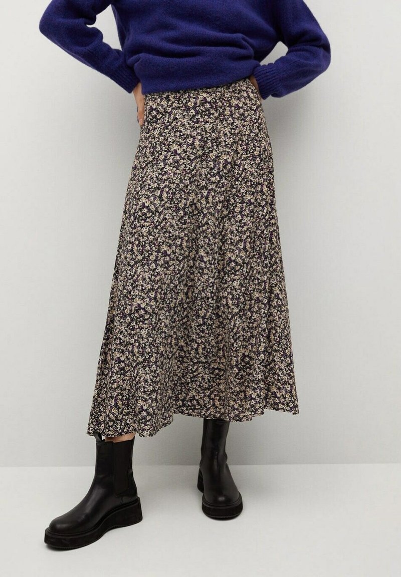 Personne portant un pull violet, une longue jupe noire à fleurs et des bottines noires, debout avec les mains sur les hanches contre un mur uni.