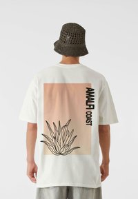 Tricou alb cu un fundal în degrade roz, text negru "AMALFI COAST" și conturul unei plante de agave. Purtați cu o pălărie de tip bucket gri, țesută.