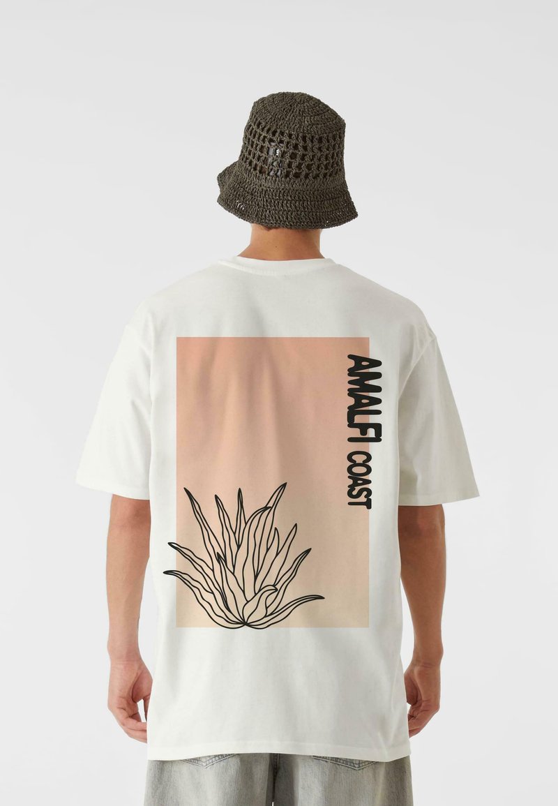 Tricou alb cu un fundal în degrade roz, text negru "AMALFI COAST" și conturul unei plante de agave. Purtați cu o pălărie de tip bucket gri, țesută.