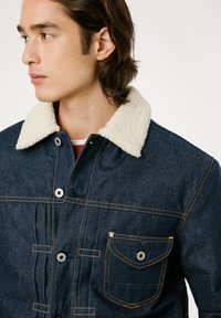Chaqueta de mezclilla oscura con un cuello de sherpa crema, botones en la parte frontal, un bolsillo en el pecho con cierre de botón y costuras naranjas en contraste.