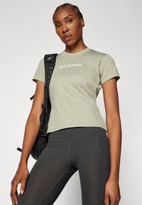 Olijfgroen overhemd met korte mouwen en een ribbeltextuur, voorzien van een wit logo, gecombineerd met donkergrijze skinny leggings en een zwarte rugzak.