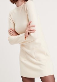 Robe-pull couleur crème en tissu tricoté doux, avec manches longues, col montant et deux poches plaquées à l'avant.