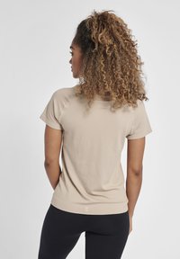 Beżowa koszulka z krótkim rękawem o fakturowanej tkaninie, okrągłym dekolcie i prostym wykończeniu. Noszona z czarnymi legginsami; zawiera mały detal z logo z tyłu.