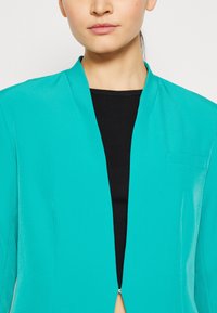 Chaqueta turquesa con una textura suave, diseño de frente abierto y un pequeño detalle de bolsillo en el lado izquierdo. Llevada sobre una blusa negra.