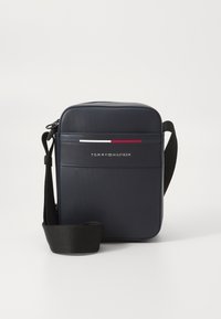Mornarska Tommy Hilfiger torbica preko tijela napravljena od teksturiranog materijala; ima crni remen, logotip na prednjoj strani i suptilne crvene i bijele dizajnerske akcentne.