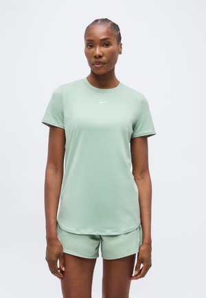 Kvinde stående vendt fremad iført en lysegrøn Nike T-shirt med korte ærmer og matchende shorts mod en ensfarvet baggrund.