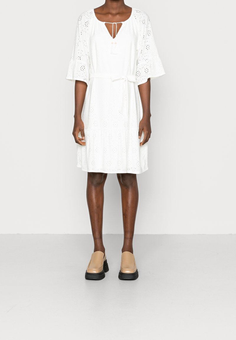 Robe en dentelle blanche avec des manches évasées, un col rond et un détail à nouer ; présente des motifs en œillets ; associée à des chaussures beige à enfiler avec des semelles noires.