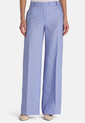 Kobieta ubrana w jasnoniebieskie spodnie z wysokim stanem i szerokimi nogawkami, w połączeniu z beżowymi butami w szpic, stojąca na tle jednolitego tła.