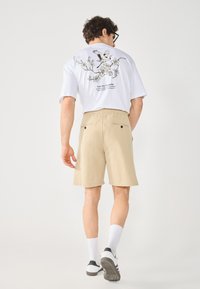 T-shirt blanc avec un motif d'oiseau noir et de fleurs au dos, associé à un short beige avec deux poches arrière et une ceinture élastique.