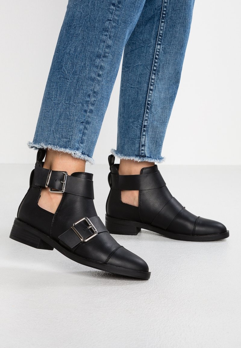 Bottes basses en cuir noir avec un design ajouré, des boucles doubles sur les côtés et un talon plat. Portées avec un jean en denim bleu délavé et usé.