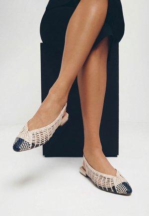 Piernas cruzadas con zapatos slingback planos tejidos en beige y azul marino con punta puntiaguda, sentadas sobre un cubo negro contra un fondo blanco.