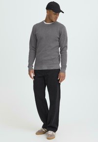 Pulover gri tricotat cu decolteu rotund, pantaloni negri lejeri și adidași gri cu accente albe și bej. Șapcă neagră completează ținuta.