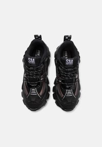 Zapatillas deportivas negras con paneles de malla, suelas de goma y detalles brillantes. Marca destacada en la lengüeta y la tira de sujeción.