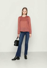 Blusa plissada cor ferrugem com mangas longas, combinada com jeans flare azul-escuro e botas pretas de tornozelo, segurando uma pequena mala preta.