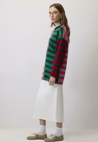 Gestreepte trui in groen, rood en roze met lange mouwen en geribbelde boorden, gecombineerd met een witte midi-jurk en beige schoenen.