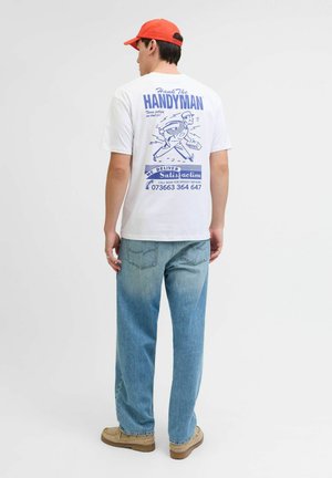 Man in een wit graphic t-shirt, lichtblauwe jeans, tan schoenen en een rode pet, staande met de rug naar de camera tegen een effen witte achtergrond.