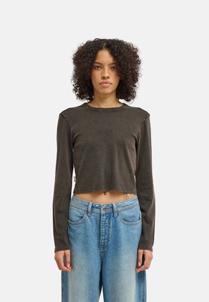 Persoon met kort, krullend haar draagt een donker lang crop-top met losse blauwe jeans en staat frontaal tegen een effen lichte achtergrond.
